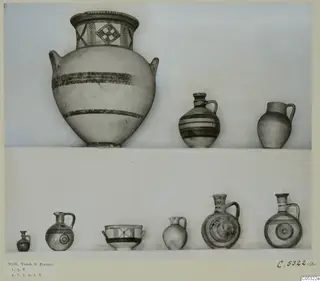 Pottery vessels from Stylli, Tomb 8, arranged on two white shelves against a plain background