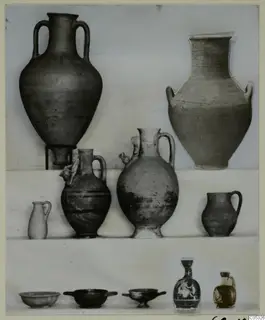 Pottery from Marion, Tomb 18: two large jars, several jugs and small bowls set out on shelves against a plain backdrop