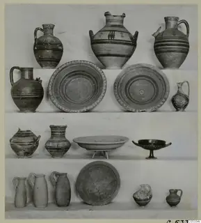 Many pottery vessels, jugs, jars, dishes and a two-handled cup, arranged in four rows on white-draped shelves.