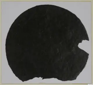Dark, corroded circular object with a broken notch on its right edge, shown on a pale card. Marion, Tomb 72.