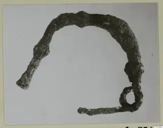 Corroded bronze object, open arc with a small loop at one end, photographed on a mounted card
