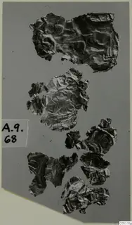 Crumpled, thin metal fragments scattered on a grey mount, with a handwritten number label at the left edge