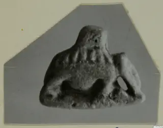 Small figure-like object with a rounded head, ridged body and pierced holes, shown on a grey card mount