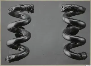 Two spiral gold objects, each with a moulded head-like terminal at the top, photographed side by side on a grey ground.
