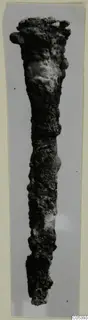 Long, heavily corroded iron tool photographed upright against a plain grey background