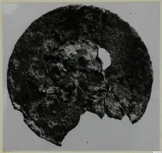 Corroded bronze disc fragment with an off-centre hole, from Marion Tomb 9, shown on a grey background.