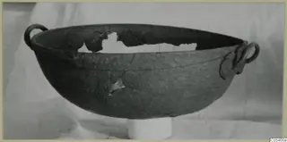 Dark, corroded metal bowl with two loop handles, shown on a white pedestal