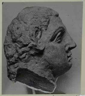 Profile photograph of a carved stone head from Mersinaki, facing right, with wavy hair and a pitted surface.