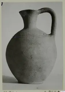 Pottery jug with a single loop handle and a narrow neck, grey-white surface with faint horizontal grooves on the shoulder.
