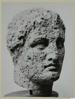 Stone head of a figure from Mersinaki, with short hair and a heavily pitted surface, photographed on a round stand.