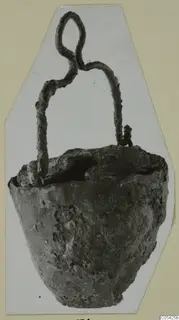 Corroded metal lamp with a looped wire handle rising from the rim, photographed alone on a pale card.