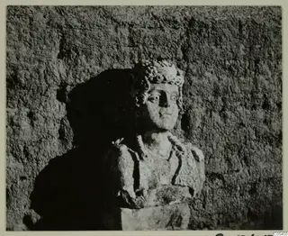 Carved stone bust of a figure with a wreath or curled headdress, set against a rough textured wall.