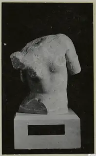 Marble sculpture of a human torso with the head and arms broken, on a white plinth against a black background.