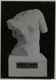 Torso of a carved pale stone statue on a white pedestal, shown against a black backdrop