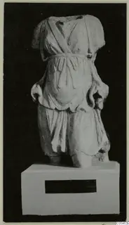 Marble sculpture of a headless standing figure in a belted tunic, shown on a plinth against a black background.