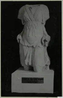 Marble sculpture of a draped standing figure, headless and armless, on a white plinth against a black background.