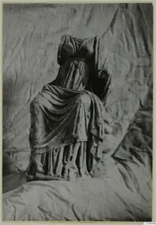Headless seated draped statue in marble, photographed against a crumpled cloth backdrop.
