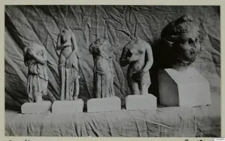 Four small standing stone figures on white plinths and a larger carved head on a block, photographed against draped cloth.