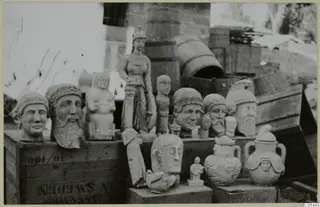 Sculpted heads, a standing statue and two lidded two-handled vessels set out on wooden crates outdoors