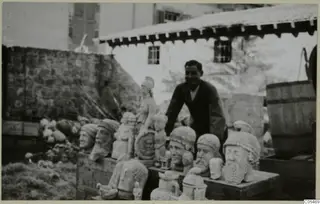 Smiling man leaning over a crate of carved stone heads and busts in an outdoor yard, with a tiled-roof building behind