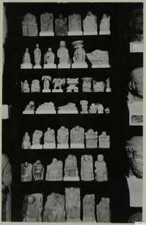 Rows of small limestone figurines and reliefs on shelves against a dark background, with large stone heads at the right edge.