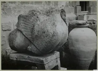 Carved stone sculpture fragment from Soli on a stone block pedestal, with a tall jar beside it.