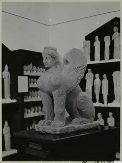 Stone sculpture of a winged creature with a human head and lion-like body, on a dark pedestal in a display room