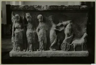 Carved pale stone relief showing four standing female figures and a wheel with an animal at right