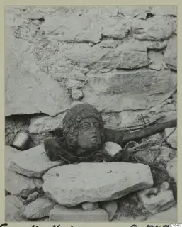 Stone head with curled hair resting among rough stones against a wall, Fasoulla-Kastros