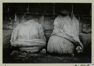 Two headless draped stone statues standing side by side against a stone wall, Arsos