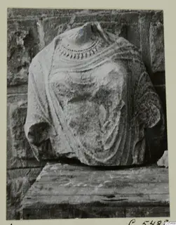 Stone statue torso without head, with folded drapery and a dotted neckline border, standing on a wooden block against a stone wall