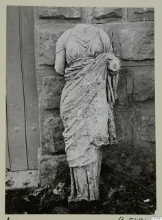 Headless draped stone statue of a standing female figure, photographed against a stone wall beside a wooden door.
