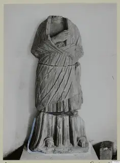 Headless stone statue of a draped standing figure from Arsos, shown against a plain wall.
