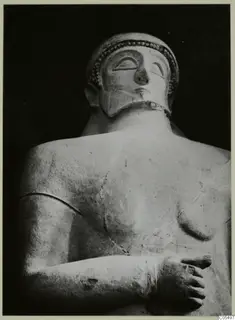 Back and side view of a bearded statue's head, shoulders and folded arm against a dark background.