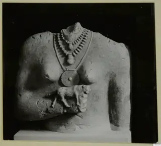 Headless limestone sculpture of a torso with a layered necklace and a small animal figure at the chest.