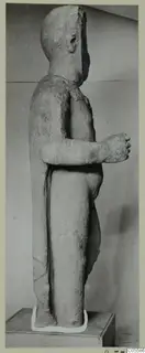 Standing limestone statue of a figure from Mersinaki, shown in side profile against a grey wall