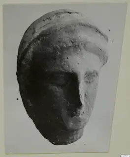 Fragment of a sculpted head from Mersinaki with a banded headdress and broken lower face, shown on a grey backdrop.