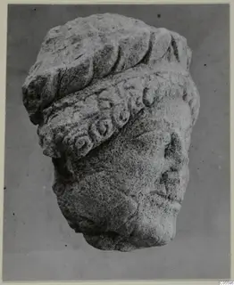 Limestone sculpture of a head wearing a thick wreath-like headdress, from Mersinaki, photographed against a plain grey background.