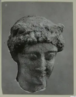 Limestone head of a figure with a rolled headdress and downcast eyes, photographed from the front against a grey backdrop