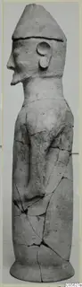 Terracotta standing figure in profile, wearing a tall conical cap, with one arm bent across the body