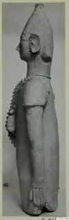 Terracotta standing figure in side view, with a tall head and serrated ridges over the shoulder and chest.