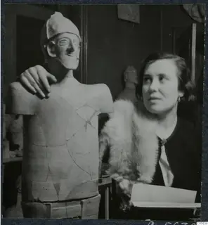Woman in a fur stole beside a reconstructed statue of a figure with a helmet-like headdress, a hand resting on its shoulder.