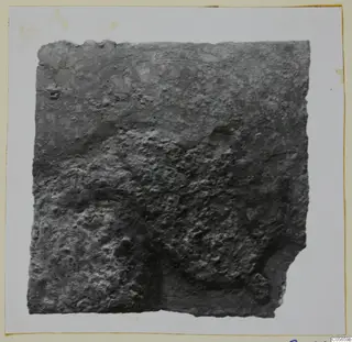 Corroded bronze fragment from Marion Tomb 60, with a raised curved pattern and broken edges, photographed on a card mount.