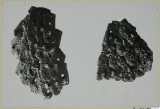 Two corroded iron fragments on a pale background, the larger one made of overlapping plates with small round holes.