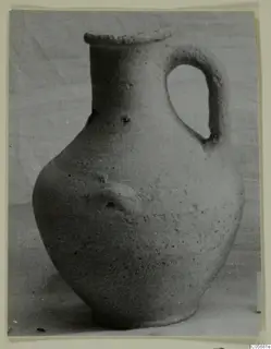 Pottery jug with a single loop handle and a small applied knob on the body, shown against a plain cloth background