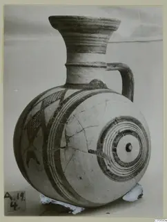 Round-bodied pottery jug with a flaring neck, a handle and painted bands and geometric patterns in dark paint on a pale surface.