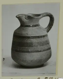Jug with a pinched spout, a loop handle and dark painted bands, shown in a photograph mounted on a card.