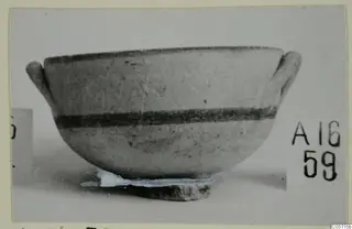Pottery bowl with two horizontal handles and dark painted bands, photographed on a stand against a grey background.