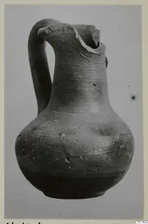 Pottery juglet with a looped handle and a broken rim, photographed against a plain grey background