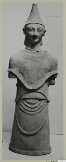Terracotta figure with a tall conical cap, short stub arms and a pleated skirt, photographed against a grey backdrop